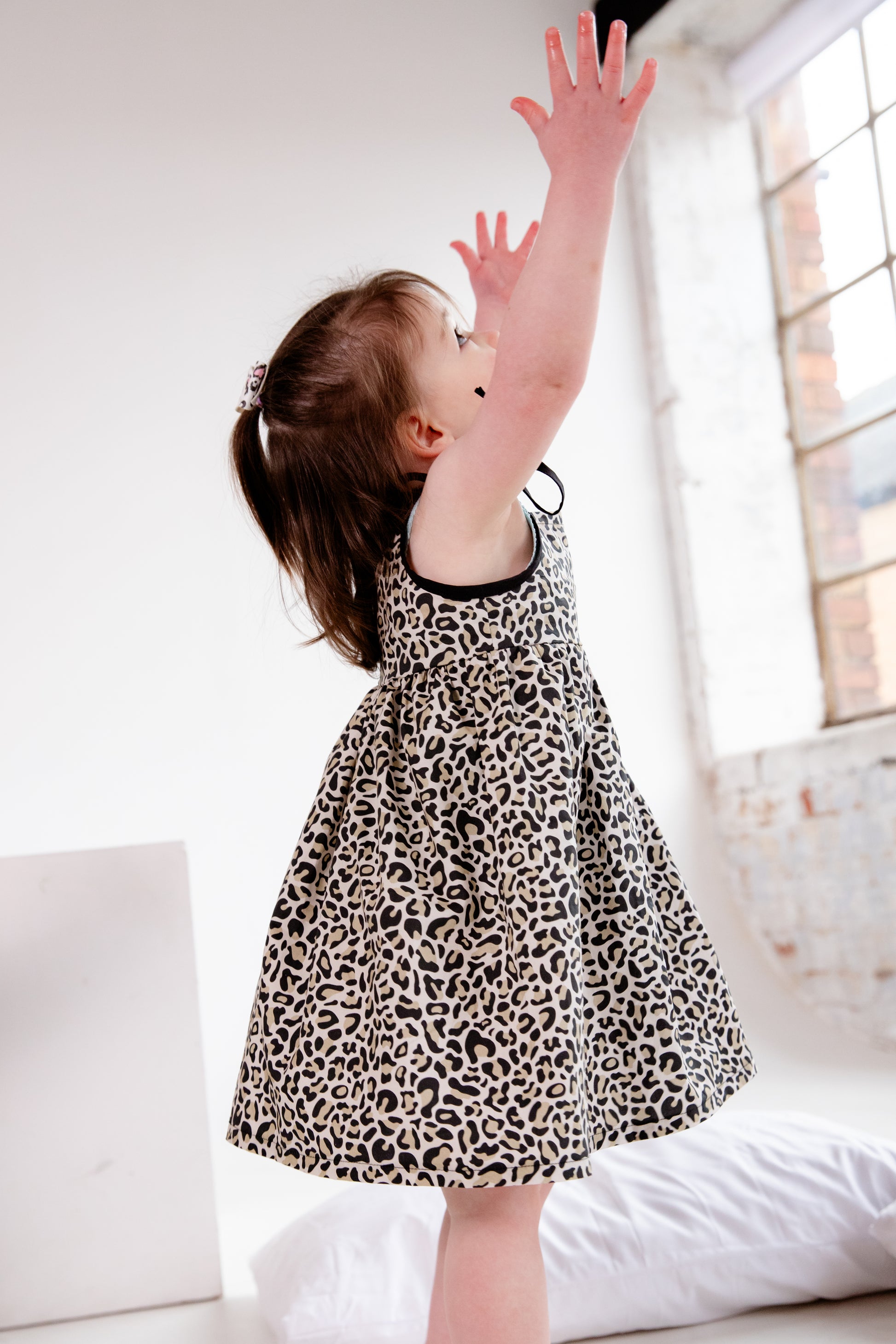 A young child wearing a handmade dress from Oxfordshire featuring a black and white leopard print pattern. The dress has a sleeveless design with delicate black ribbon ties on the shoulders, a gathered waistline, and a flowing skirt. The fabric has a soft yet structured appearance, complementing the child’s playful movement. The child is barefoot, standing on a textured cream-colored couch, adding to the cozy and natural feel of the image.