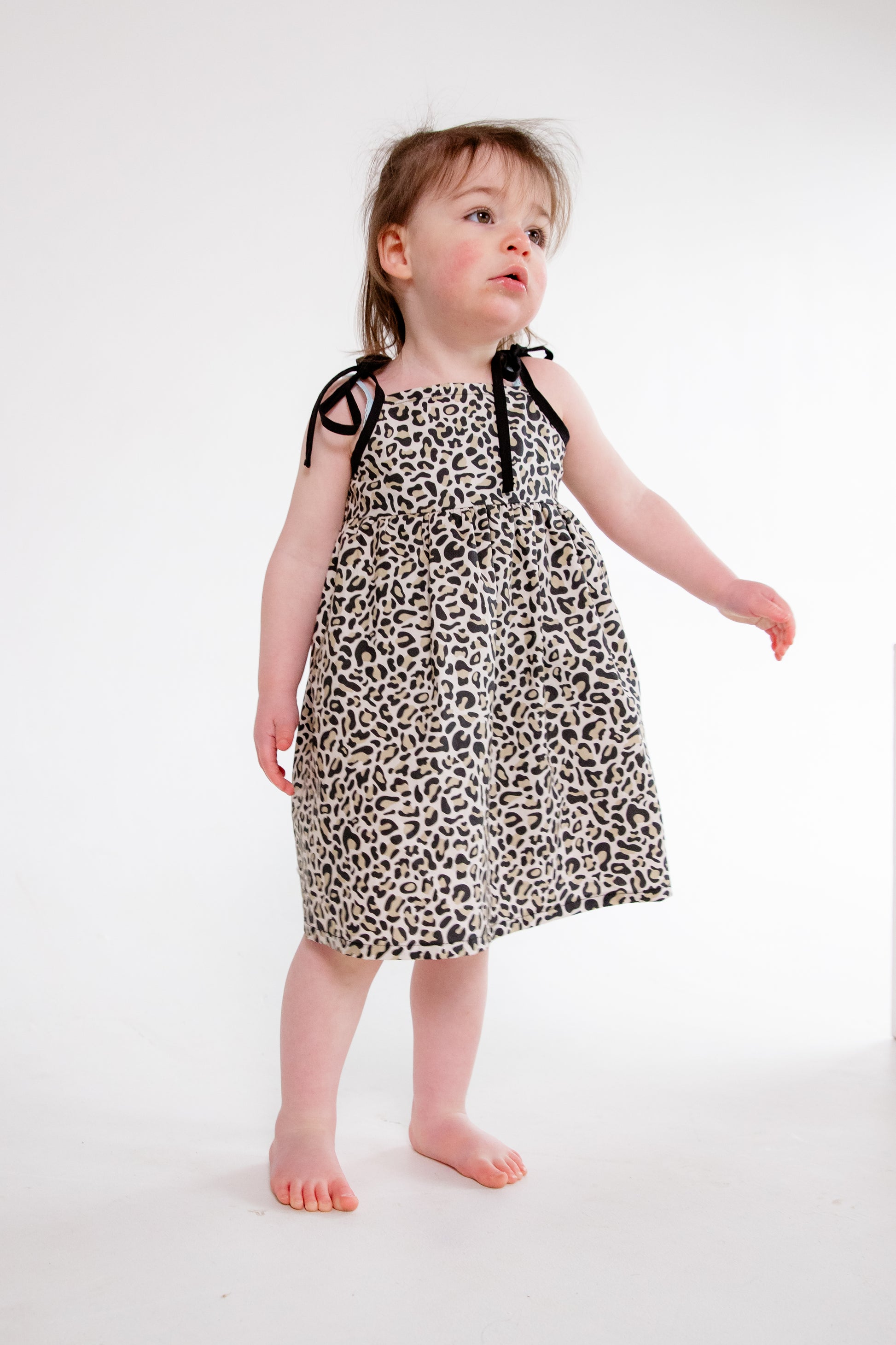 A young child wearing a handmade dress from Oxfordshire featuring a black and white leopard print pattern. The dress has a sleeveless design with delicate black ribbon ties on the shoulders, a gathered waistline, and a flowing skirt. The fabric has a soft yet structured appearance, complementing the child’s playful movement. The child is barefoot, standing on a textured cream-colored couch, adding to the cozy and natural feel of the image.