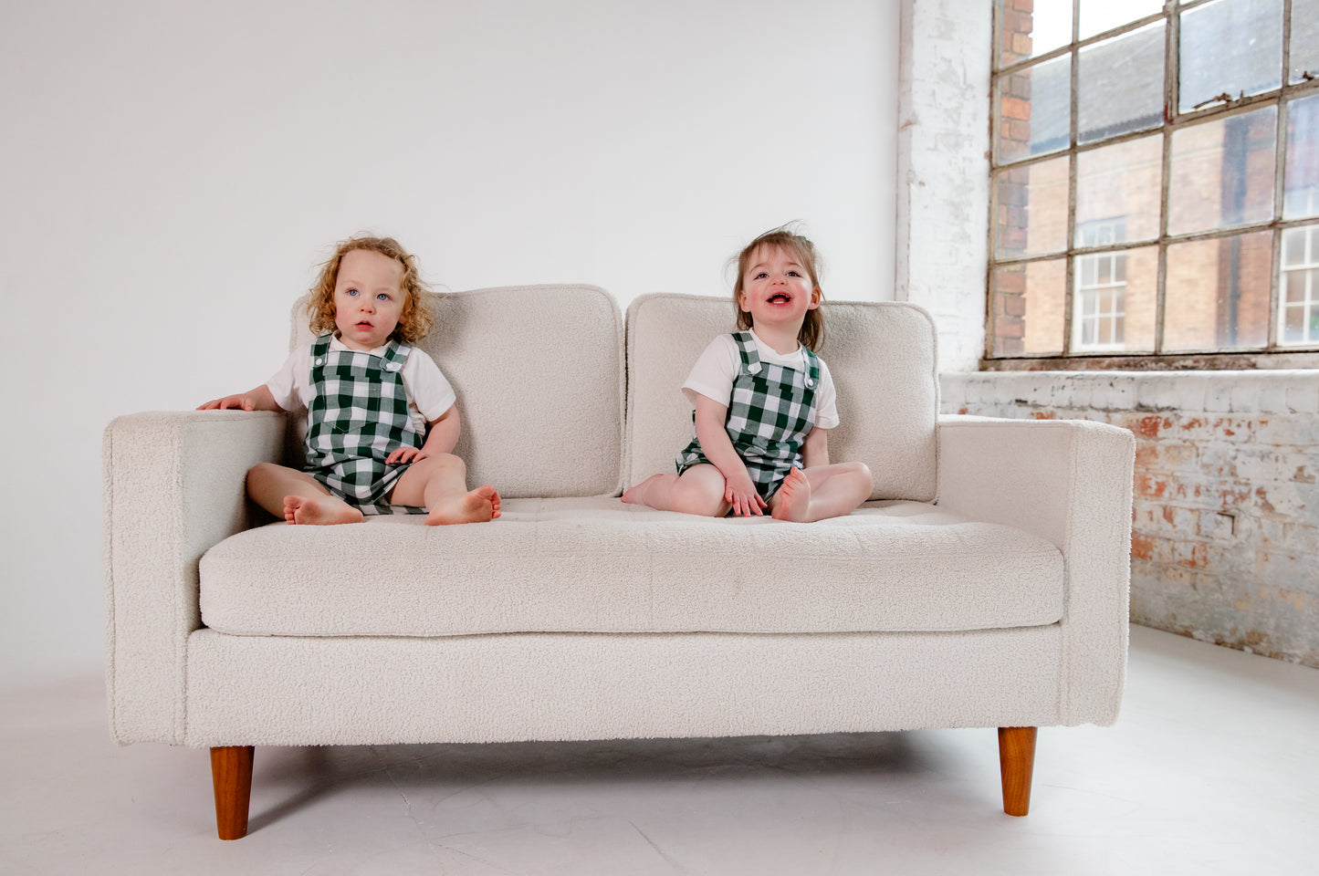 A toddler wearing a handmade green and white gingham romper crafted from cotton fabric in West Oxfordshire. The romper features adjustable shoulder straps with button fastenings, a front pocket, and a loose, comfortable fit. It is paired with a simple white short-sleeved T-shirt. The outfit showcases a timeless, classic design, perfect for everyday wear or special occasions.