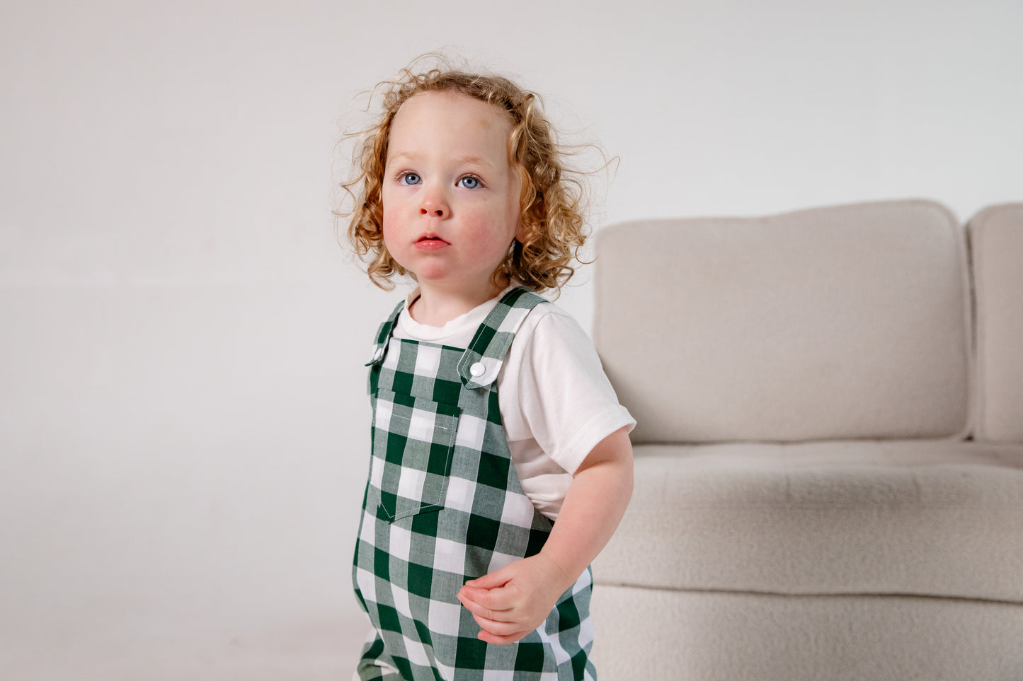 A toddler wearing a handmade green and white gingham romper crafted from cotton fabric in West Oxfordshire. The romper features adjustable shoulder straps with button fastenings, a front pocket, and a loose, comfortable fit. It is paired with a simple white short-sleeved T-shirt. The outfit showcases a timeless, classic design, perfect for everyday wear or special occasions.