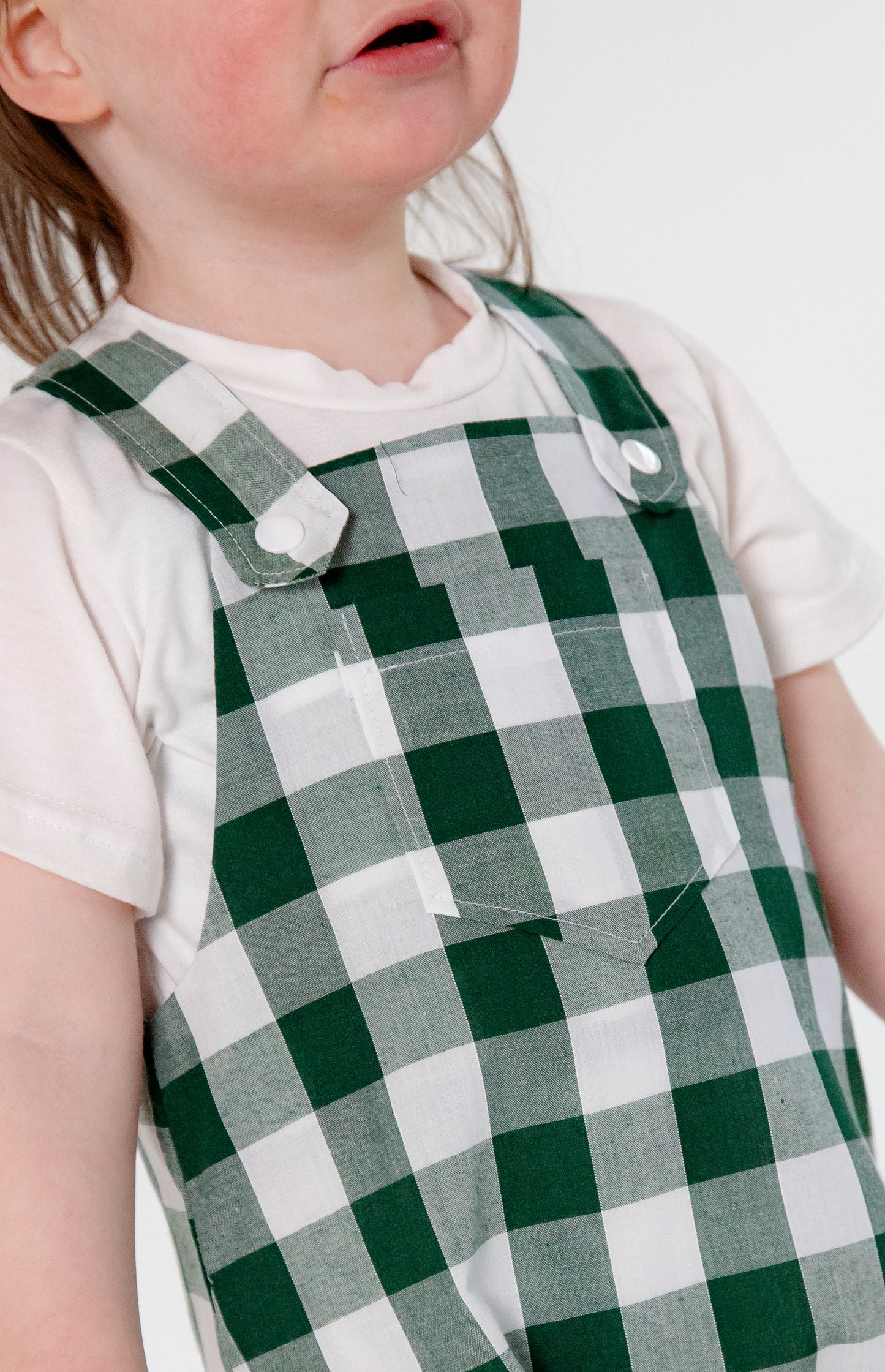 A toddler wearing a handmade green and white gingham romper crafted from cotton fabric in West Oxfordshire. The romper features adjustable shoulder straps with button fastenings, a front pocket, and a loose, comfortable fit. It is paired with a simple white short-sleeved T-shirt. The outfit showcases a timeless, classic design, perfect for everyday wear or special occasions.