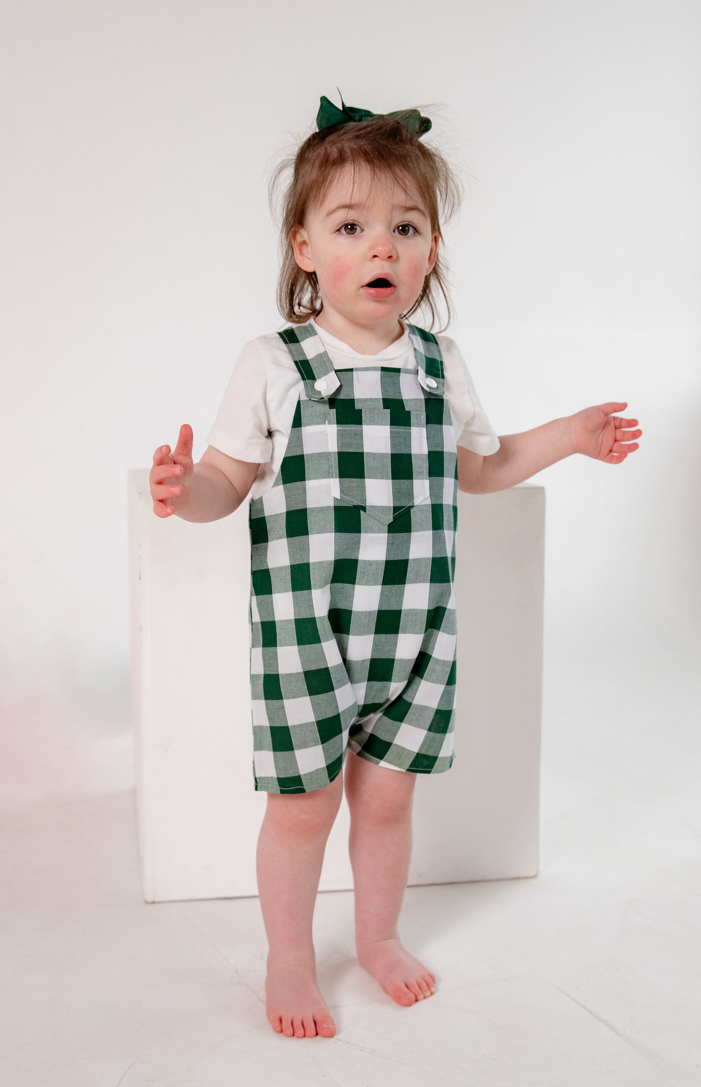 A toddler wearing a handmade green and white gingham romper crafted from cotton fabric in West Oxfordshire. The romper features adjustable shoulder straps with button fastenings, a front pocket, and a loose, comfortable fit. It is paired with a simple white short-sleeved T-shirt. The outfit showcases a timeless, classic design, perfect for everyday wear or special occasions.