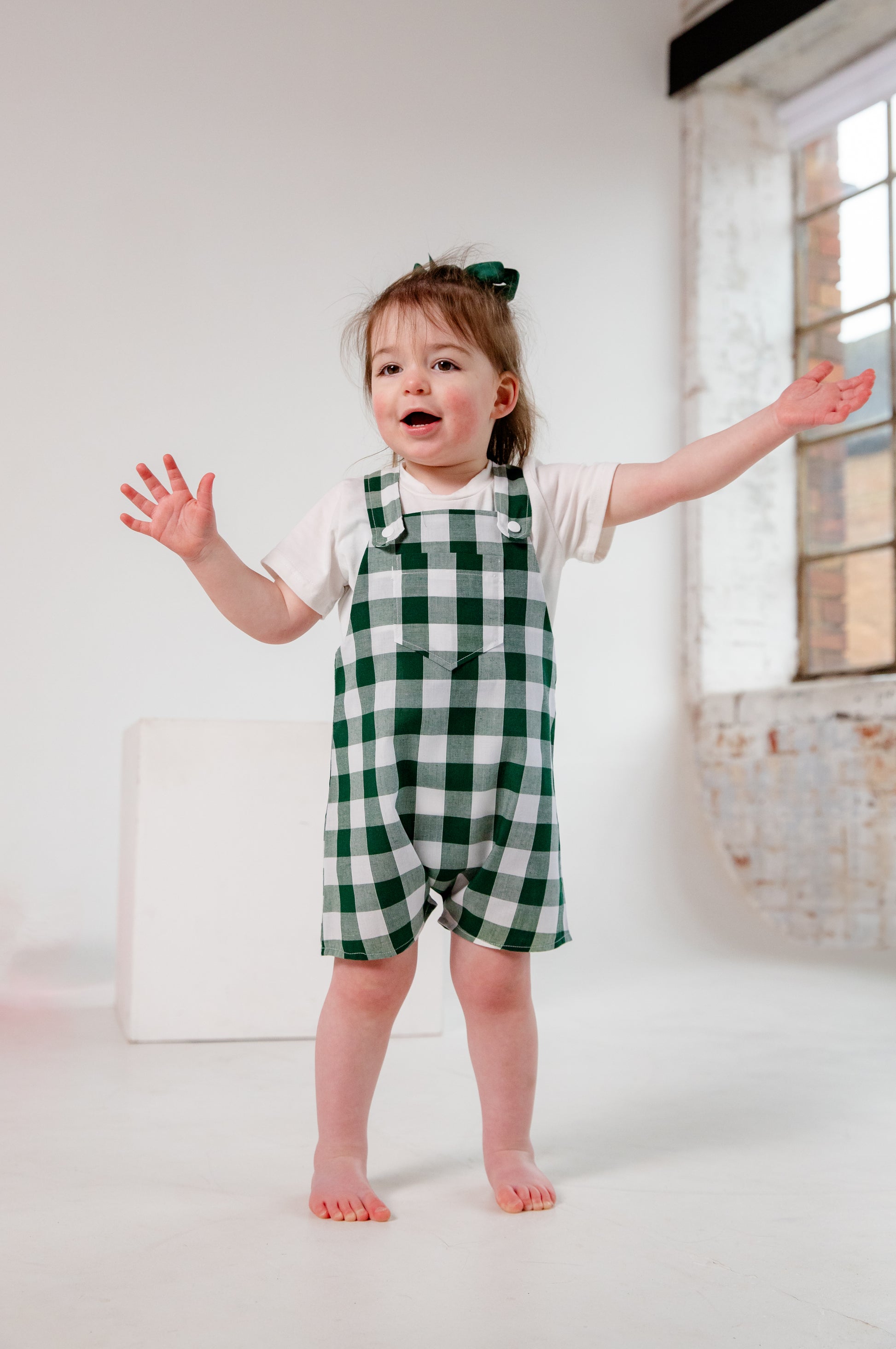 A toddler wearing a handmade green and white gingham romper crafted from cotton fabric in West Oxfordshire. The romper features adjustable shoulder straps with button fastenings, a front pocket, and a loose, comfortable fit. It is paired with a simple white short-sleeved T-shirt. The outfit showcases a timeless, classic design, perfect for everyday wear or special occasions.