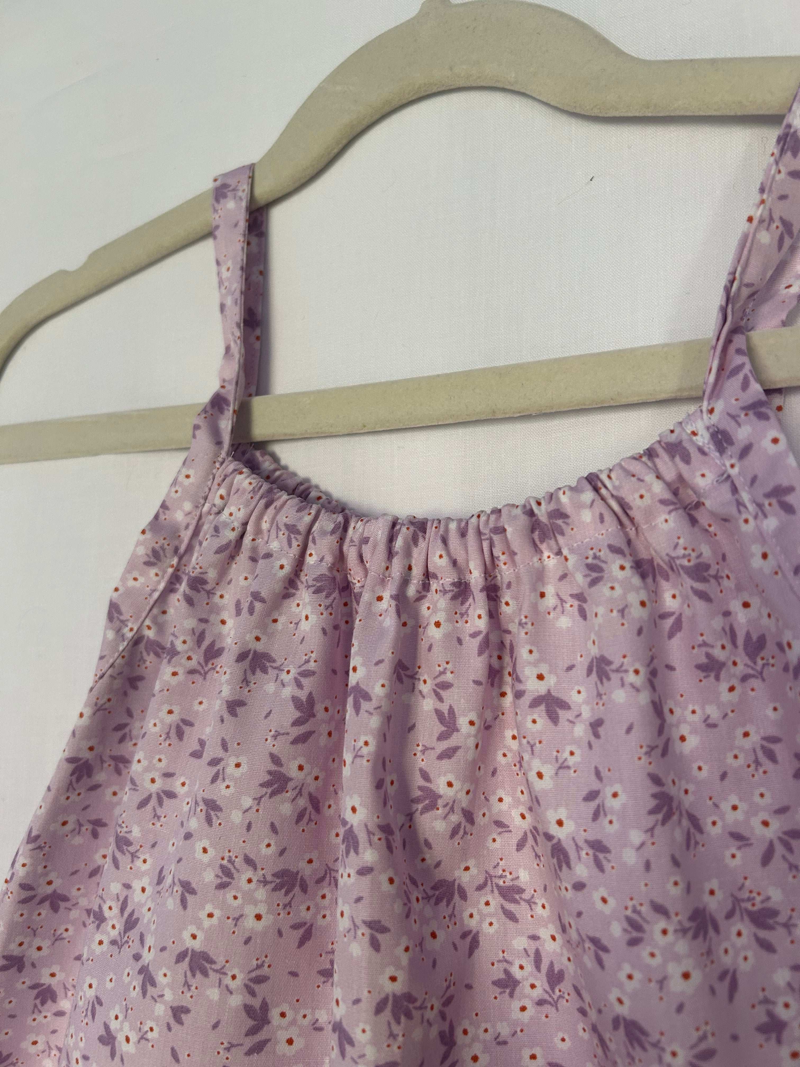 A young child wearing a handmade floral print romper. The romper has a lilac flower print on soft, lightweight fabric. It has thin, shoulder straps and ruched neckline, relaxed, comfortable fit. 