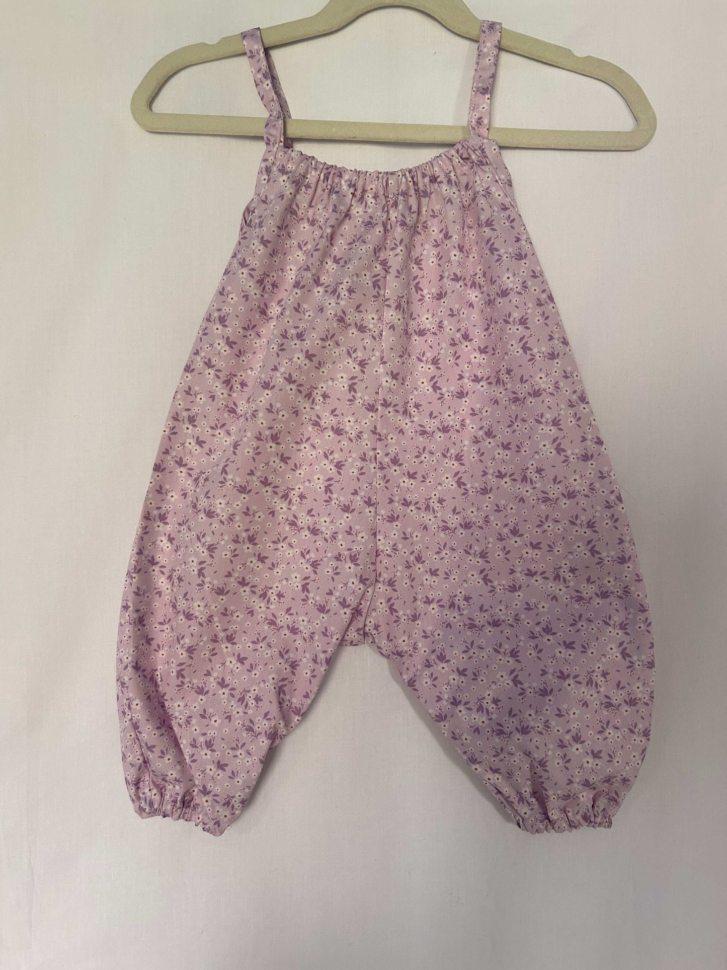 A young child wearing a handmade floral print romper. The romper has a lilac flower print on soft, lightweight fabric. It has thin, shoulder straps and ruched neckline, relaxed, comfortable fit. 