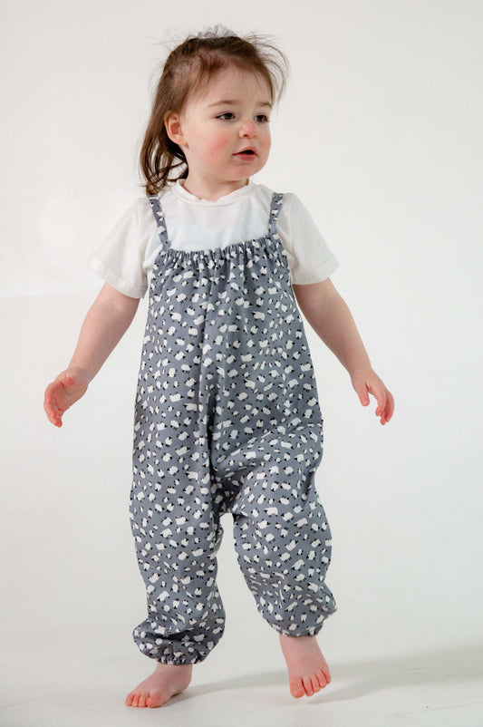A young child wearing a handmade sheep romper. The romper has a delicate grey and white sheep print on soft, lightweight fabric. It has thin, shoulder straps and ruched neckline, relaxed, comfortable fit. 