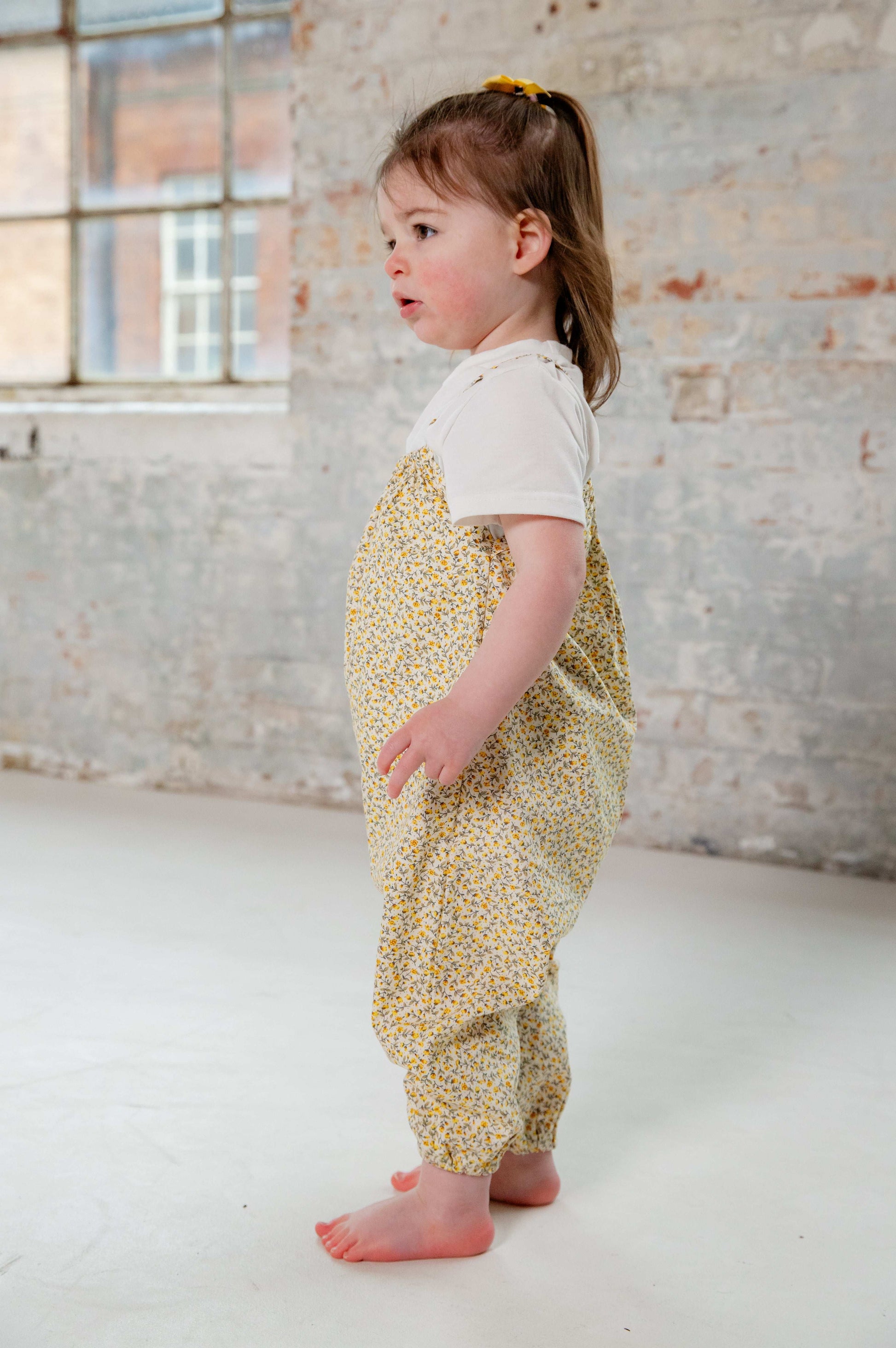 A young child wearing a handmade floral romper. The romper has a delicate yellow and white ditsy floral print on soft, lightweight fabric. It has thin, shoulder straps and ruched neckline, relaxed, comfortable fit.