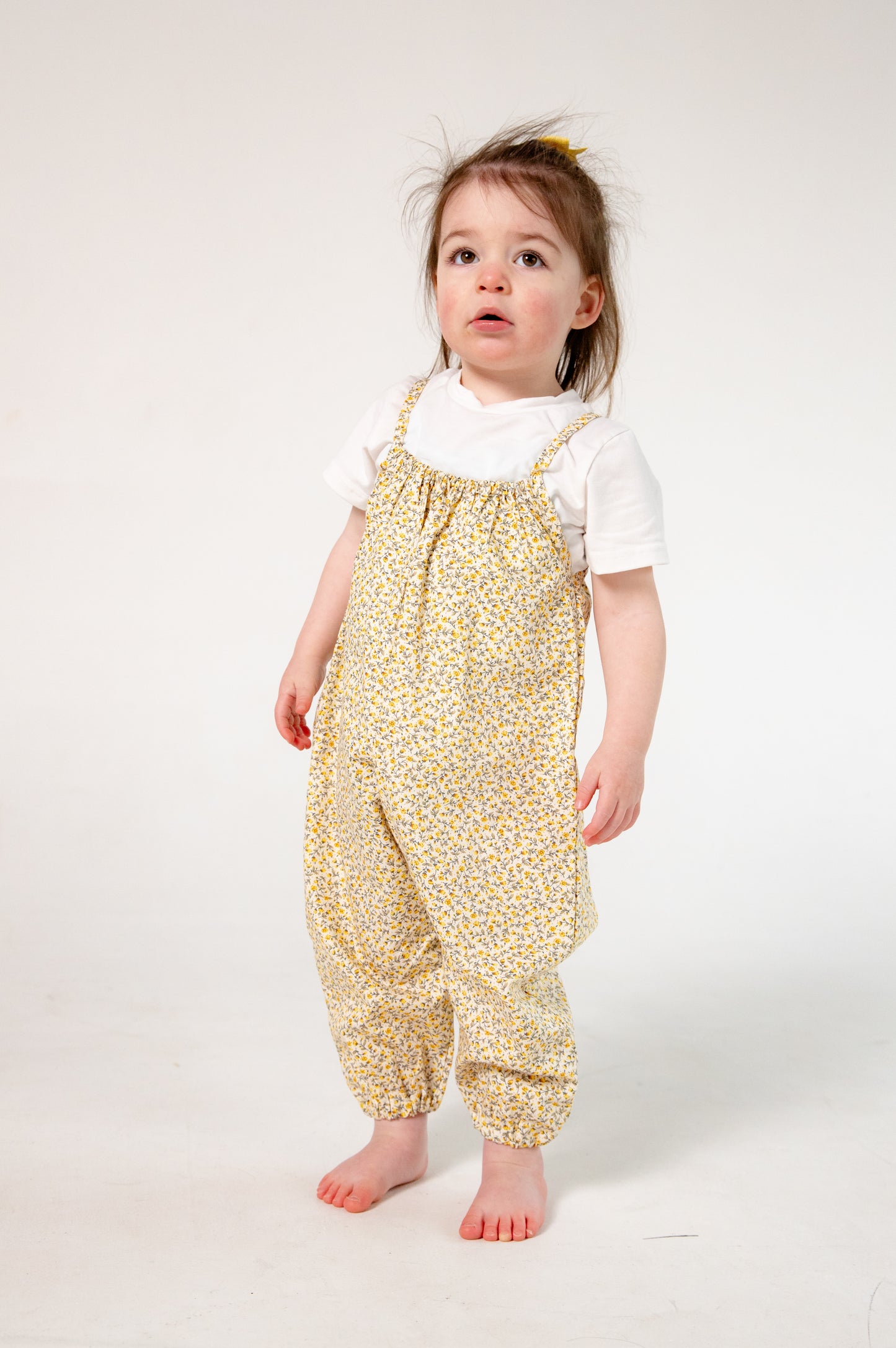 A young child wearing a handmade floral romper. The romper has a delicate yellow and white ditsy floral print on soft, lightweight fabric. It has thin, shoulder straps and ruched neckline, relaxed, comfortable fit.