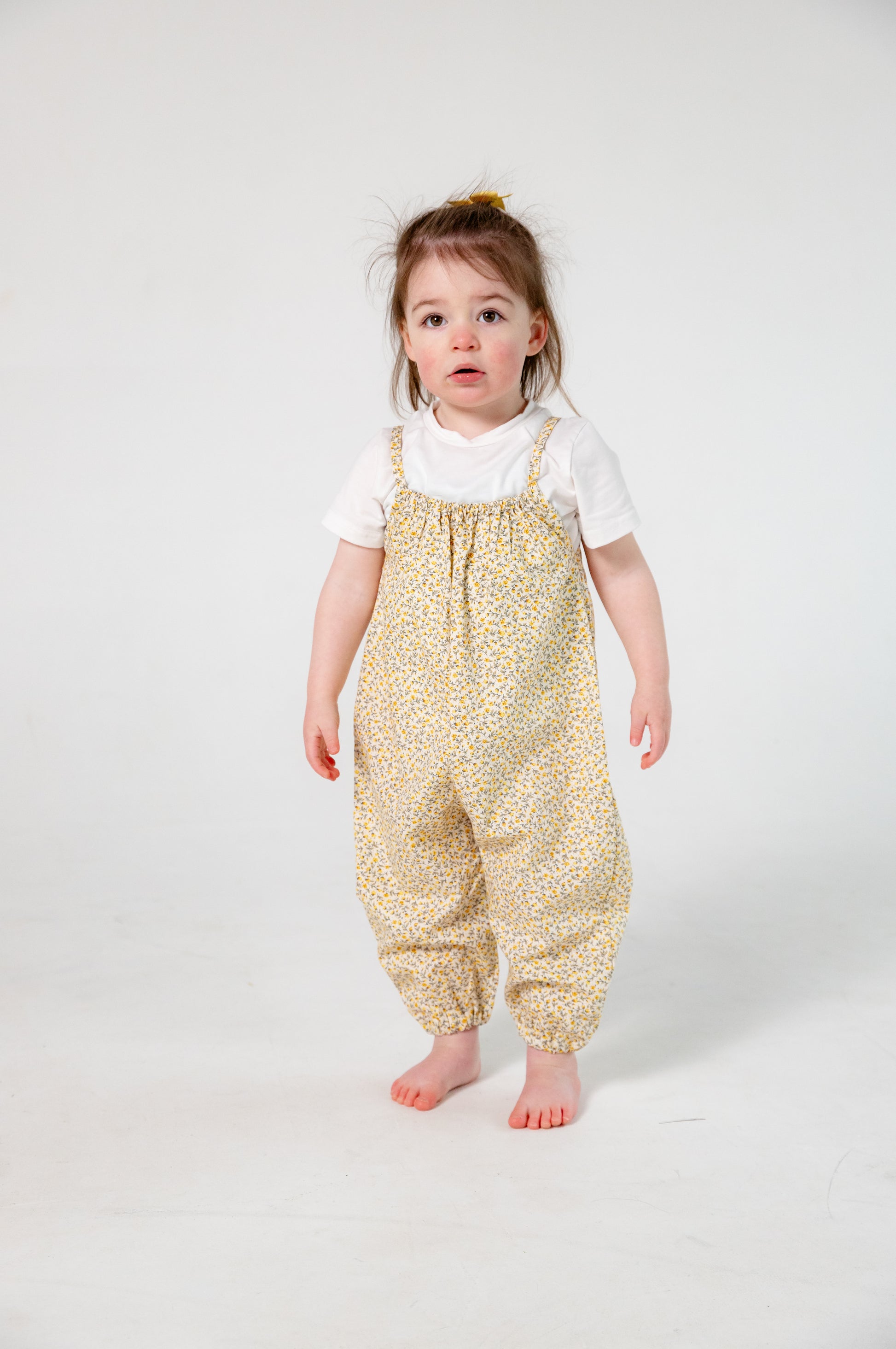 A young child wearing a handmade floral romper. The romper has a delicate yellow and white ditsy floral print on soft, lightweight fabric. It has thin, shoulder straps and ruched neckline, relaxed, comfortable fit.